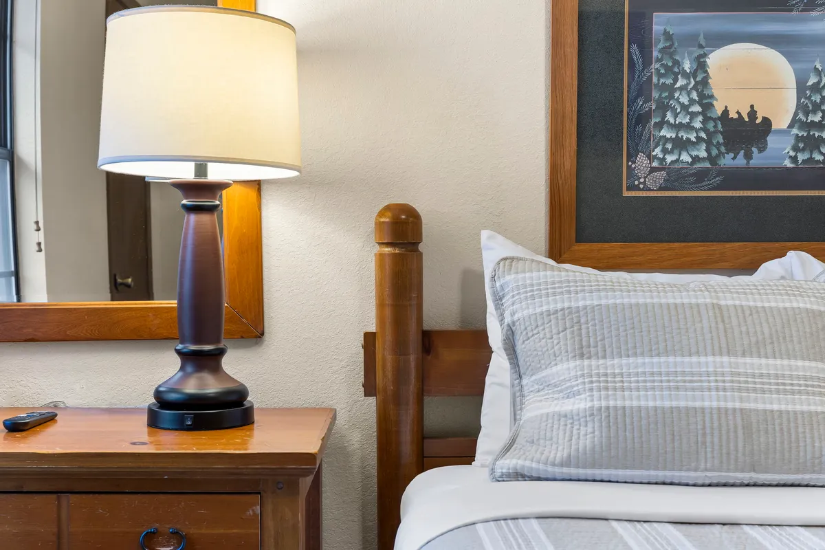 A close-up view of a cozy bed topped with luxurious linens, accompanied by a modern lamp and tasteful artwork on the wall. The scene exudes warmth and offers a comfortable, inviting nook for rest.