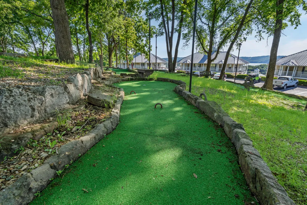 A cheerful exterior view of a mini-golf course with bright green artificial turf, rock formations, and surrounding trees. The course layout encourages group games and social interaction in a playful, natural setting.