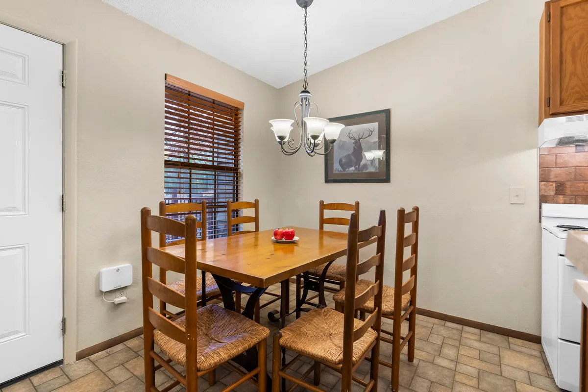 A spacious wooden dining table sits under a glowing chandelier, framed by a window with blinds and tasteful wall art. The setting is intimate and inviting, perfect for family meals, casual breakfasts, or special gatherings. The overall mood is warm and celebratory, capturing the essence of shared experiences around the table.
