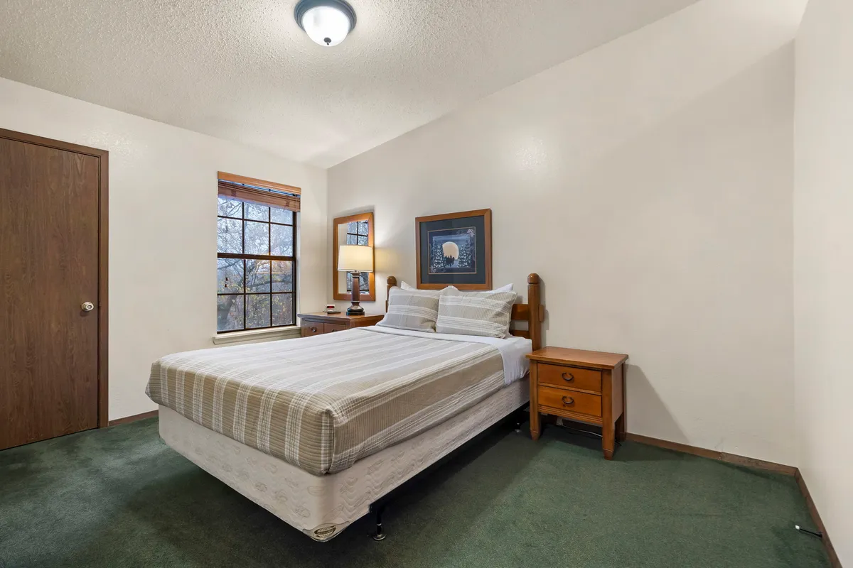 A serene bedroom featuring a queen bed with crisp linens, soft lighting from a bedside lamp, windows letting in natural light, a nightstand, and soothing wall art. The space feels quiet and inviting, ideal for relaxation.