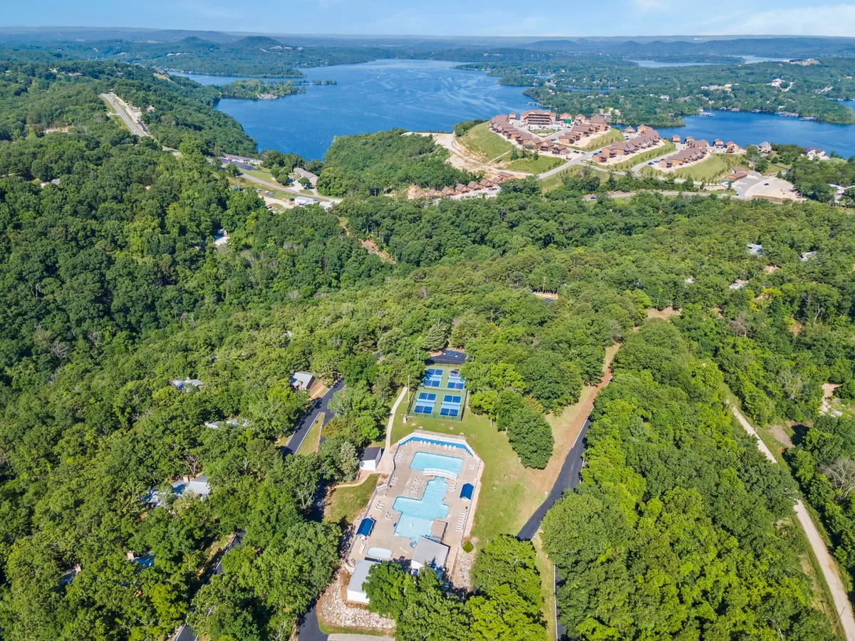 This image displays a serene outdoor swimming pool surrounded by lush greenery, with a scenic lake and residential complex visible in the background. Lounge chairs line the poolside, suggesting a space perfect for summer relaxation, social gatherings, and enjoying the beautiful Ozarks setting.