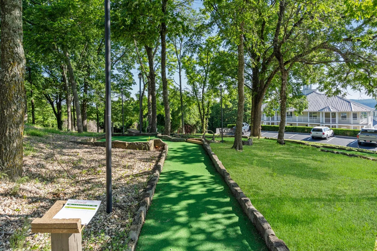 A vibrant mini-golf course winds through lush greenery, with playful landscaping and a nearby building in view. The course is clearly designed for families, with space for groups to gather and play among nature.