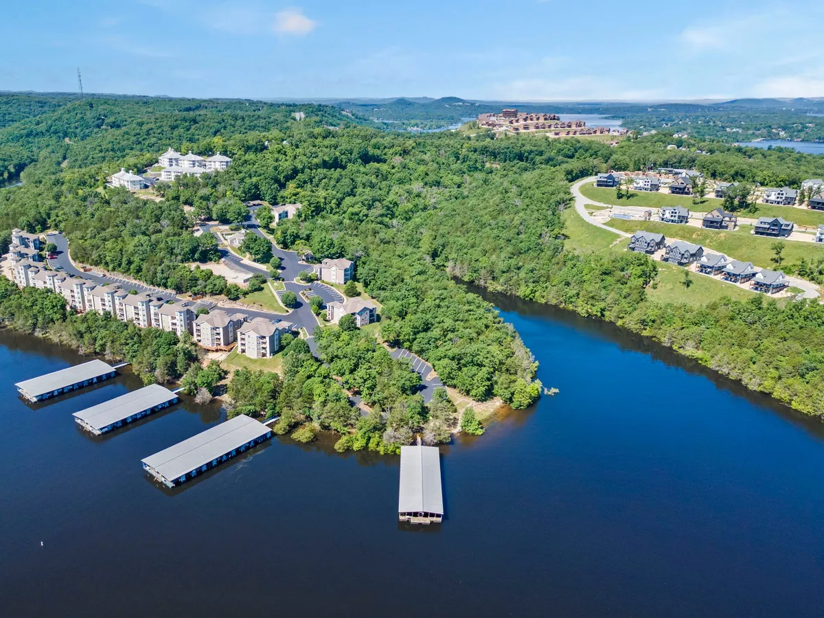 A wide waterfront scene featuring calm lake waters, a dock extending over the surface, nearby green hills, and residential buildings set among trees. The mood is tranquil, inviting guests to engage in boating, hiking, and nature walks.