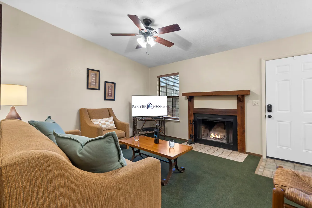 The image showcases a warm, inviting chalet living room featuring a glowing fireplace as the focal point, flanked by a comfortable sofa set and a rustic coffee table. A television is mounted for entertainment, and the room is styled for cozy gatherings and relaxation, perfect for unwinding after a day at Table Rock Lake.