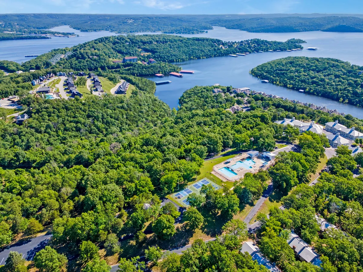 An exterior scene featuring a sparkling swimming pool in the foreground, with tennis courts and lush greenery around it. In the background, a panoramic waterfront vista hints at access to Table Rock Lake. The space is lively and recreational, inviting group activities like swimming, tennis, boating, and hiking.